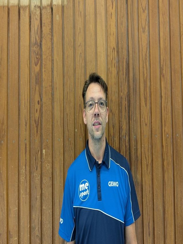 Portrait eines Mannes mit Brille, der ein blaues Sportpoloshirt vor einer Holzwand trägt.