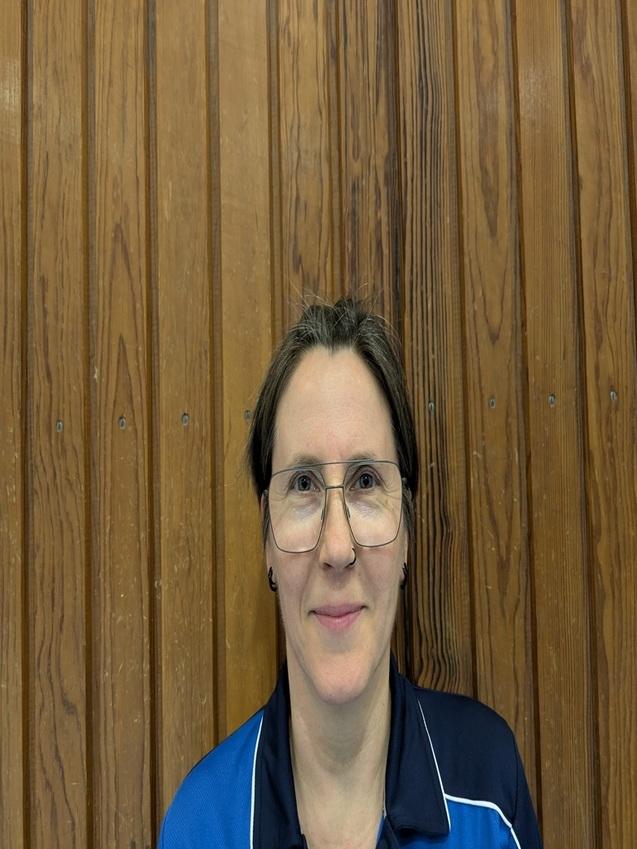 Porträt einer lächelnden Frau mit Brille, die ein sportliches Oberteil auf einem Hintergrund aus Holz trägt.