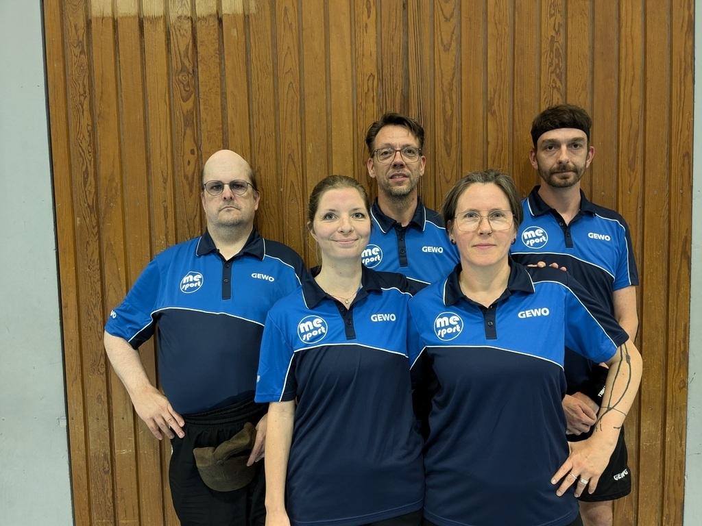 Fünf Personen in blauen Sportshirts stehen vor einer Holzwand und lächeln in die Kamera.