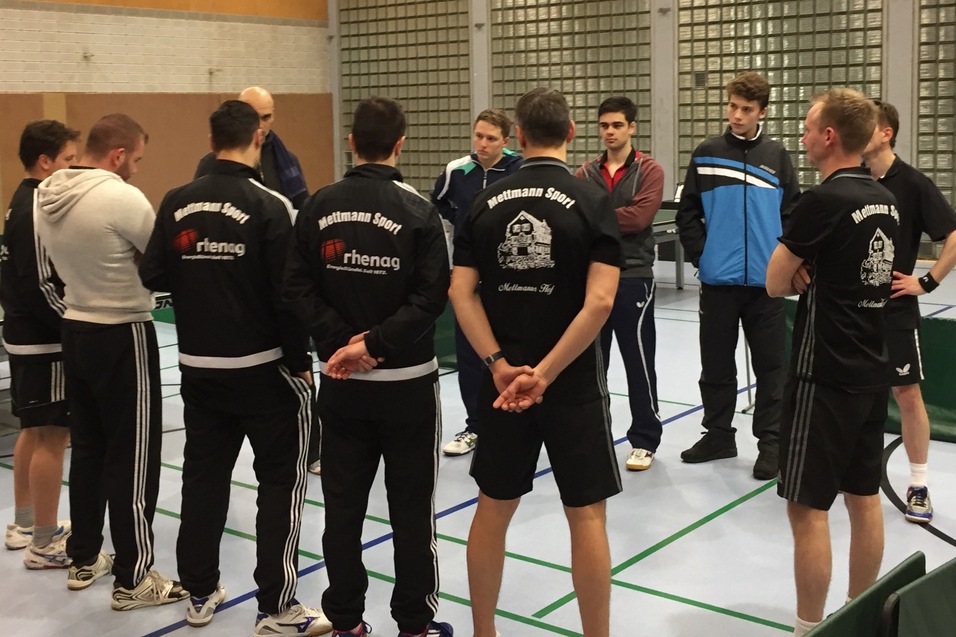 Gruppe von Tischtennisspielern in Sportkleidung bei einer Besprechung in einer Sporthalle. Tischtennisplatten im Hintergrund.