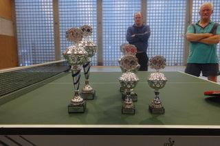 Tischtennis-Trophäen stehen auf einer grünen Tischtennisplatte, im Hintergrund zwei Männer beobachten die Szene.