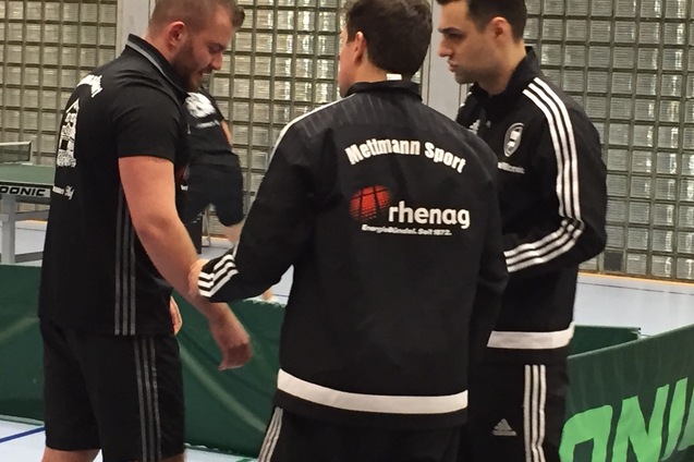 Drei Männer in Sportjacken stehen an einem Tischtennistisch und sprechen miteinander.