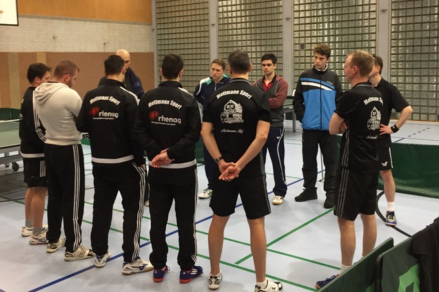 Gruppe von Tischtennisspielern in Sportkleidung bei einer Besprechung in einer Sporthalle. Tischtennisplatten im Hintergrund.