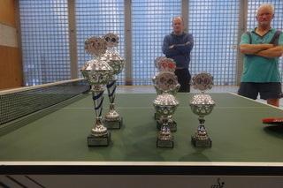 Tischtennis-Trophäen stehen auf einer grünen Tischtennisplatte, im Hintergrund zwei Männer beobachten die Szene.