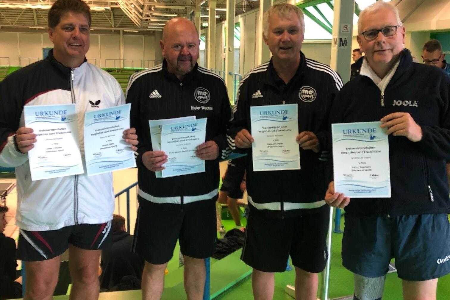 Vier Männer halten Urkunden in einer Sporthalle mit grünem Bodenbelag. Im Hintergrund sind weitere Sportler zu sehen.