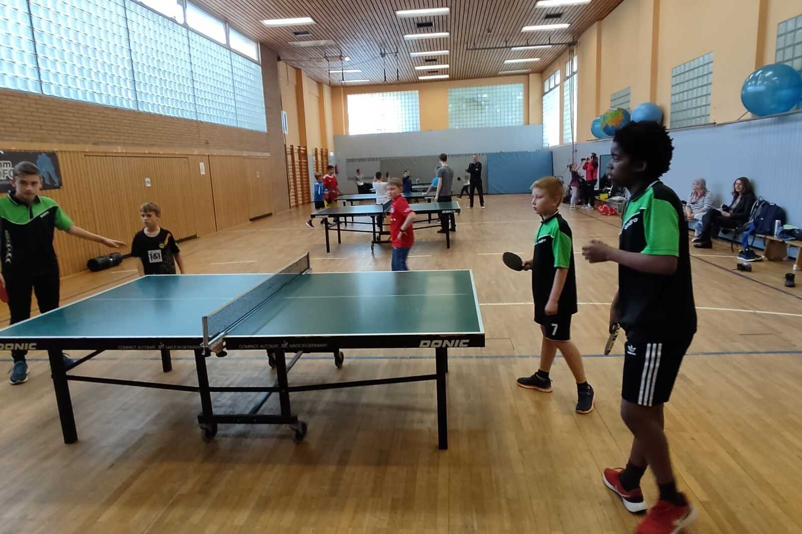 Kinder spielen Tischtennis in einer Sporthalle, umgeben von weiteren Tischen und Zuschauern.