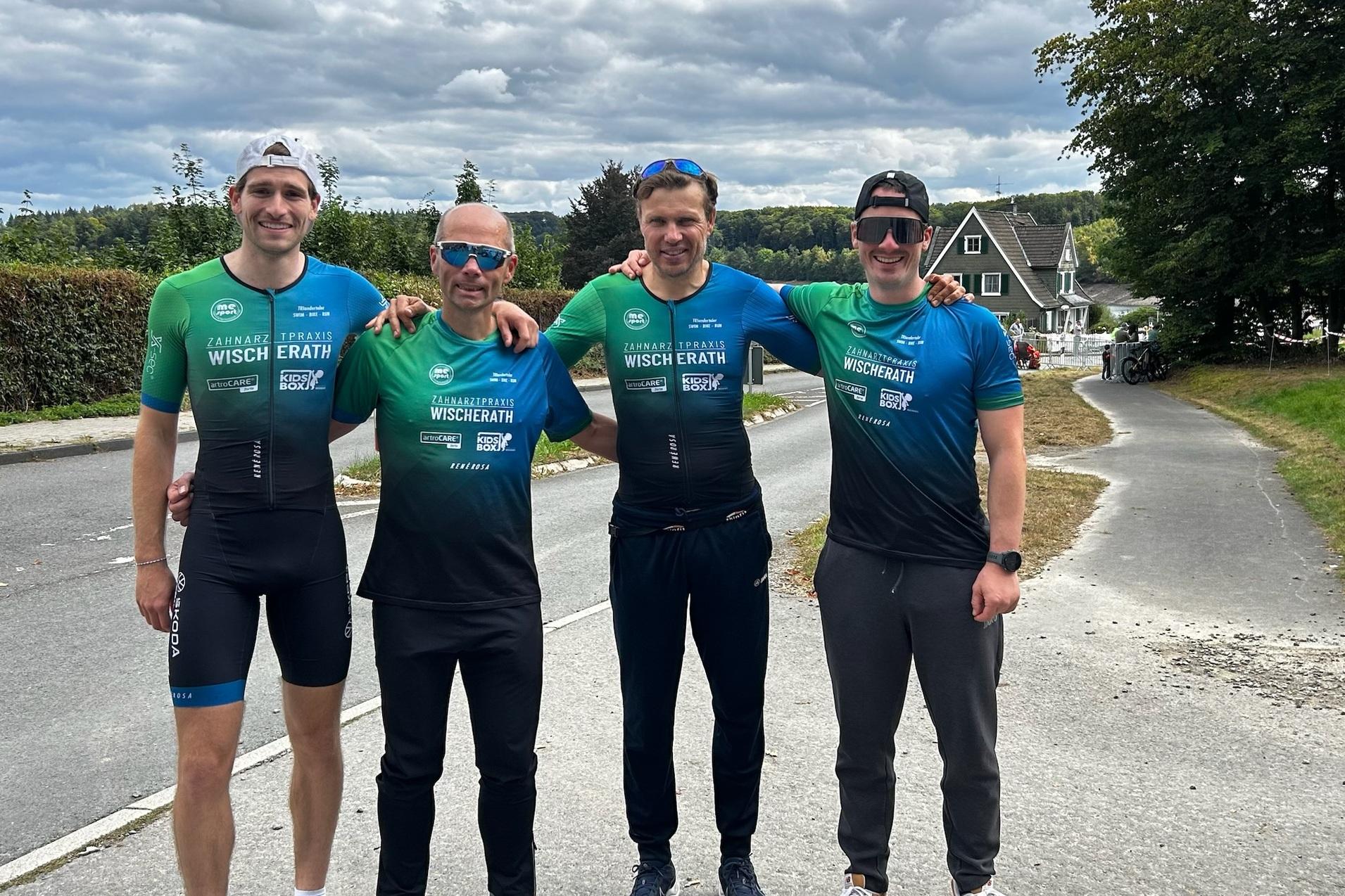 Vier M&auml;nner in Sportbekleidung stehen l&auml;chelnd an einer Stra&szlig;e, umgeben von B&auml;umen und H&auml;usern.