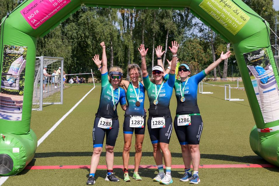 Vier Sportlerinnen feiern unter einem grünen Siegertor nach einem Wettkampf, jede trägt eine Medaille und hebt die Hände.