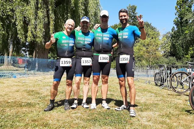 Vier Sportler in Wettkampfanzügen posieren lächelnd auf einer Wiese, umgeben von Fahrrädern und Absperrgittern.