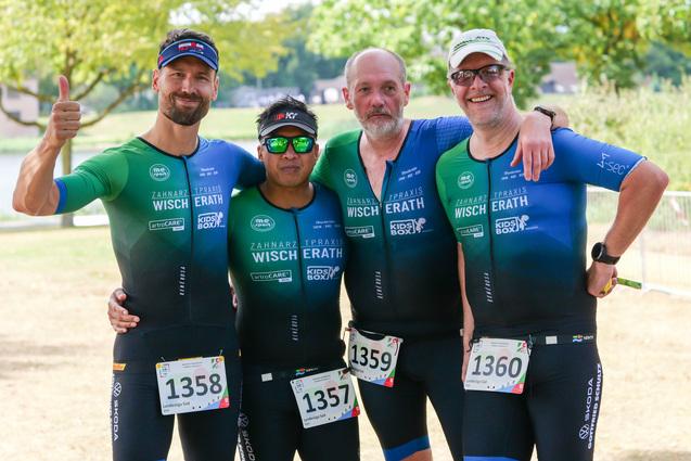 Vier Männer in Sportanzügen posieren lächelnd am Ufer eines Sees, mit Startnummern auf der Brust.