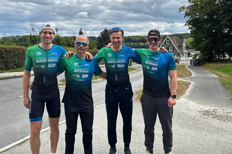 Vier Männer in Sportbekleidung stehen lächelnd an einer Straße, umgeben von Bäumen und Häusern.