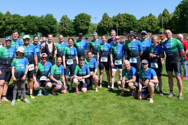 Gruppenfoto von Athleten in blauen und grünen Trikots auf einer Wiese, umgeben von Zuschauern und Bäumen.