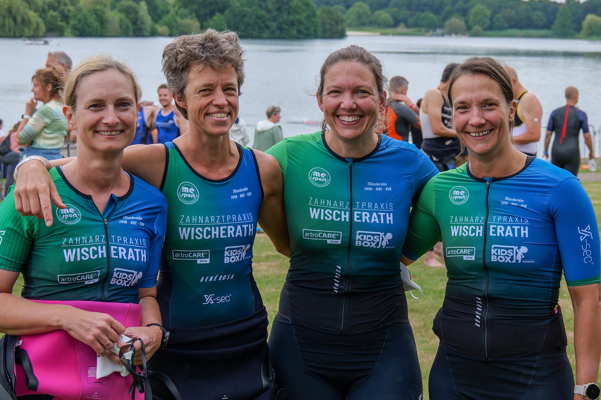 Vier Frauen in Sportanz&uuml;gen stehen l&auml;chelnd am Ufer eines Sees, umgeben von Zuschauern und Teilnehmern eines Wettkampfs.