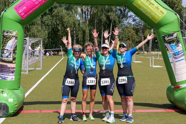 Vier Sportlerinnen feiern unter einem grünen Siegertor nach einem Wettkampf, jede trägt eine Medaille und hebt die Hände.