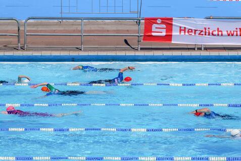 Sechs Schwimmer trainieren in einem Freibad mit Bahnmarkierungen und einer Werbebande im Hintergrund.