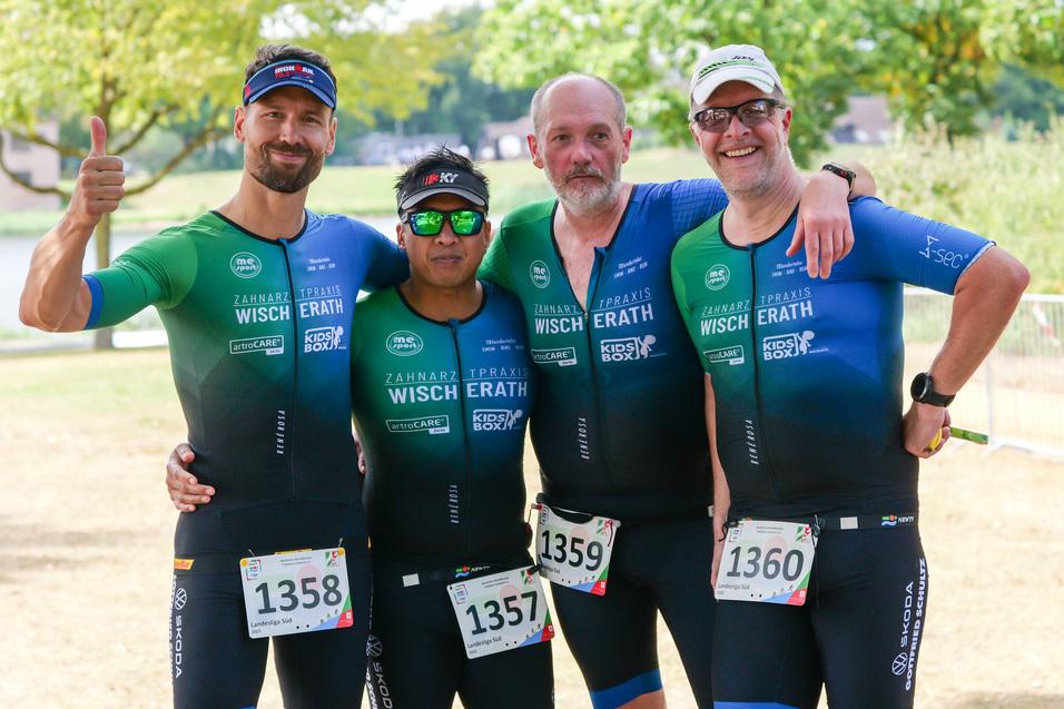 Vier Männer in Sportanzügen posieren lächelnd am Ufer eines Sees, mit Startnummern auf der Brust.