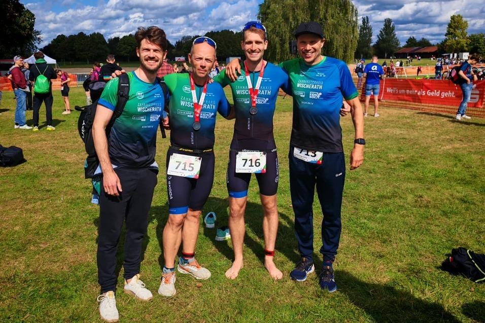 Vier Männer in Triathlonbekleidung posieren lächelnd auf einer Wiese vor einem Wettkampf, mit Medaille und Startnummern.