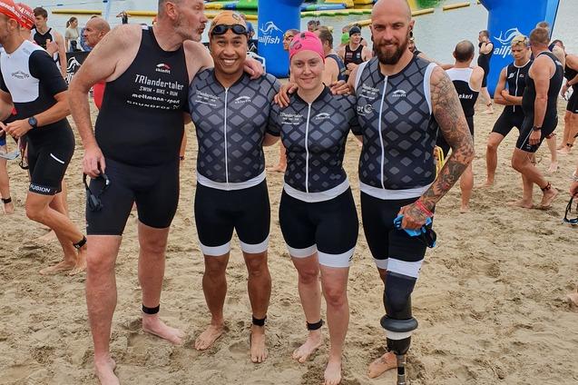 Vier Triathleten in Sportanzügen stehen am Sandstrand vor einem Stausee, mit einem blauen Zielbogen im Hintergrund.