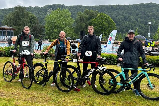 Vier Männer mit Fahrrädern stehen auf einer Wiese, im Hintergrund eine Schwimmhalle und grüne Hügel unter bewölktem Himmel.