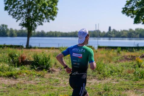 Läufer in sportlicher Kleidung joggt entlang eines Ufers mit Wasser und Bäumen im Hintergrund.