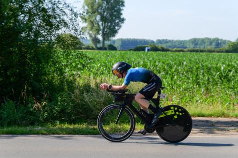 Radfahrer in aerodynamischer Position auf einem Zeitfahrrad, unterwegs auf einer Landstraße neben einem Getreidefeld.