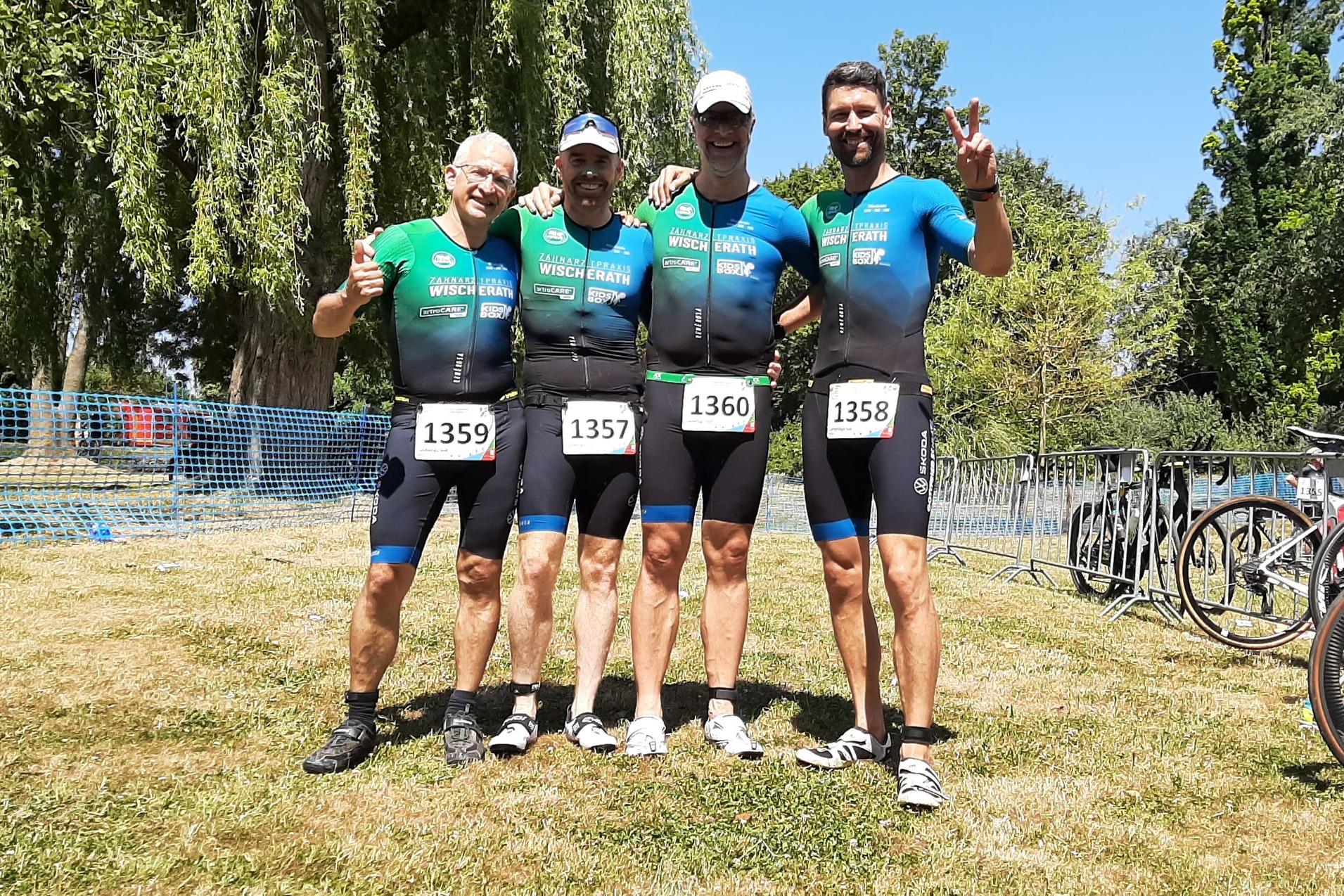 Vier Sportler in Wettkampfanz&uuml;gen posieren l&auml;chelnd auf einer Wiese, umgeben von Fahrr&auml;dern und Absperrgittern.