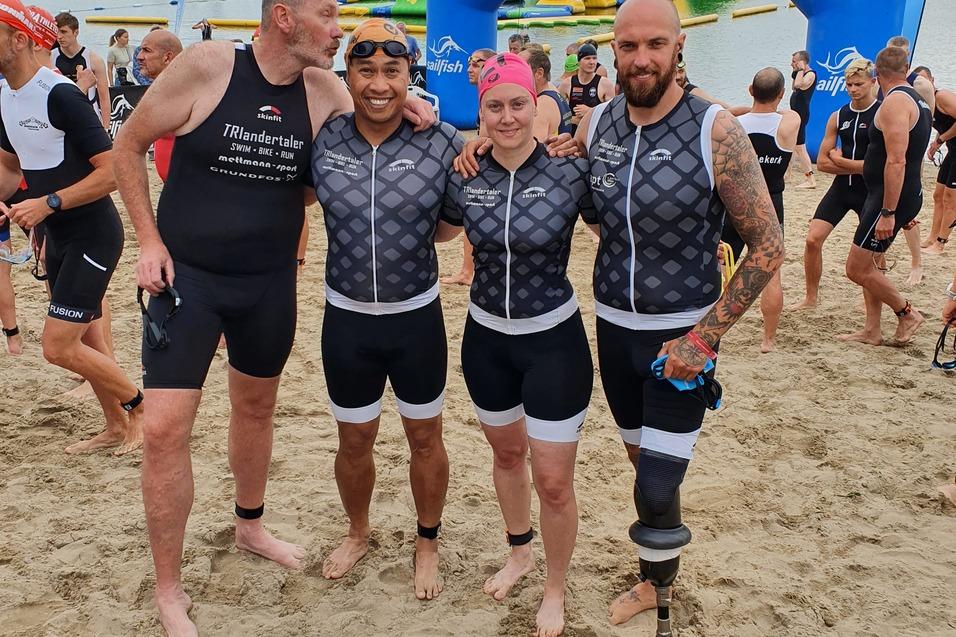 Vier Triathleten in Sportanzügen stehen am Sandstrand vor einem Stausee, mit einem blauen Zielbogen im Hintergrund.