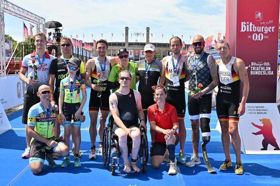 Gruppe von Triathleten mit Medaillen und Blumen auf dem Siegerpodest, Sonnenlicht, blauem Boden und Werbebanner im Hintergrund.