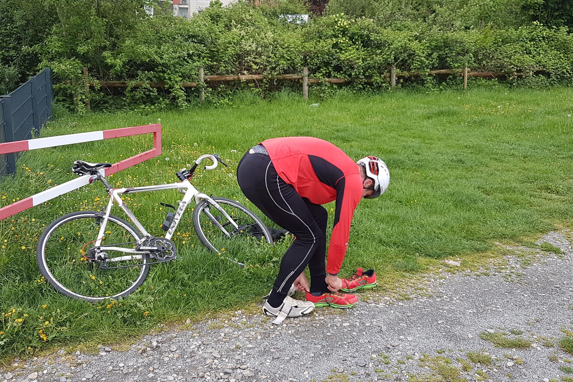Sportler in roter Bekleidung zieht sich Schuhe aus und an, während ein Fahrrad neben ihm auf dem Gras steht.