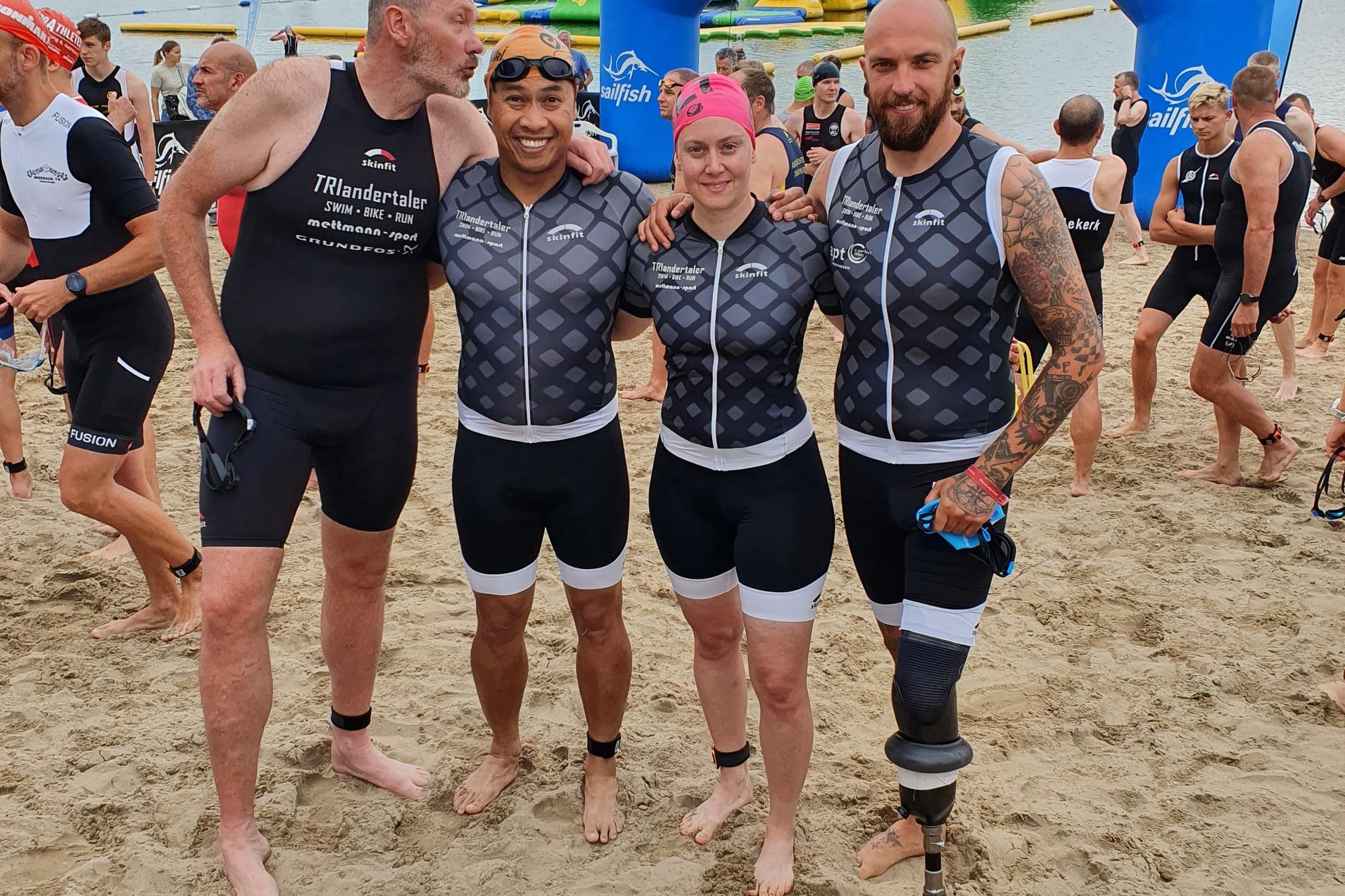 Vier Triathleten in Sportanzügen stehen am Sandstrand vor einem Stausee, mit einem blauen Zielbogen im Hintergrund.