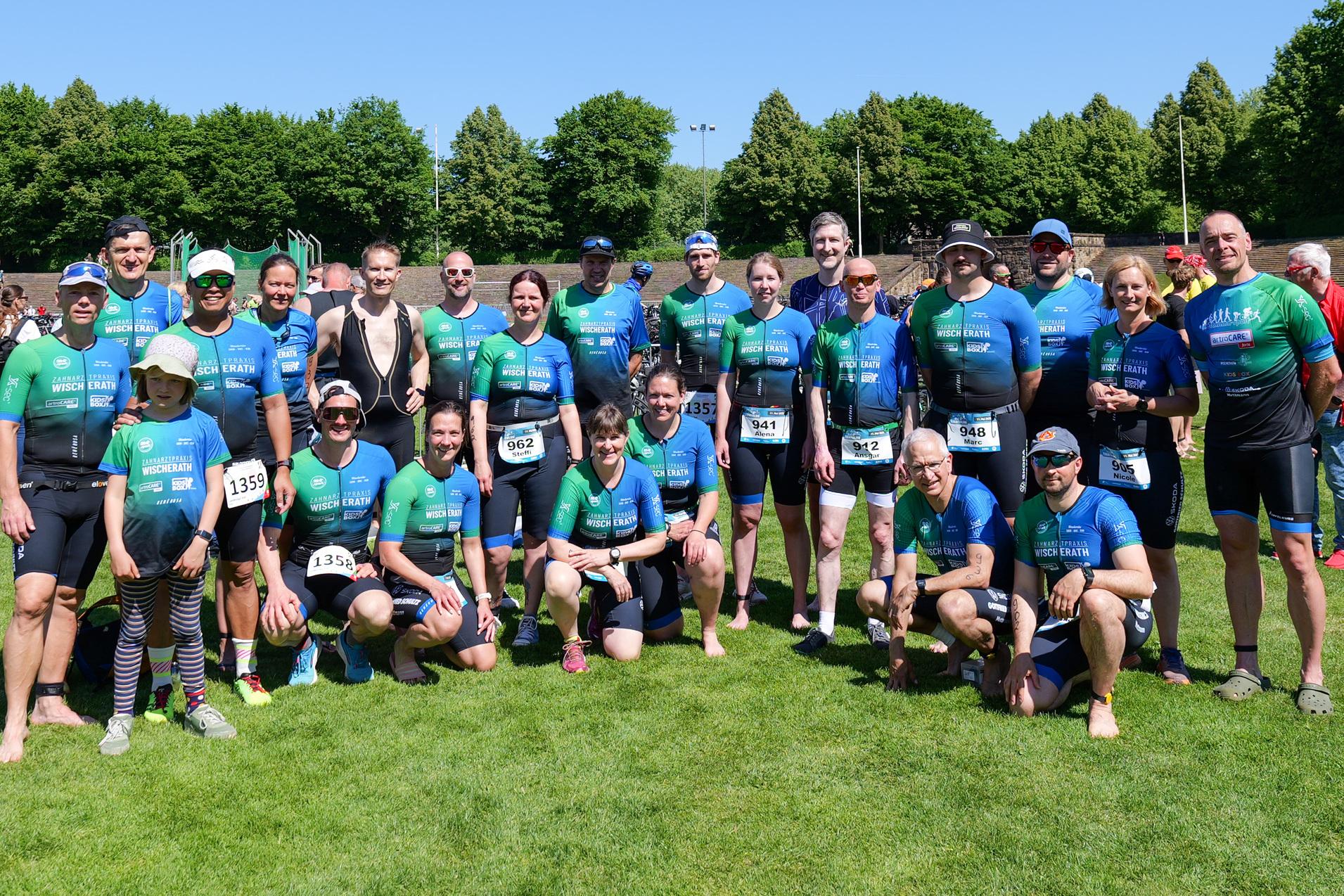 Gruppenfoto von Athleten in blauen und grünen Trikots auf einer Wiese, umgeben von Zuschauern und Bäumen.