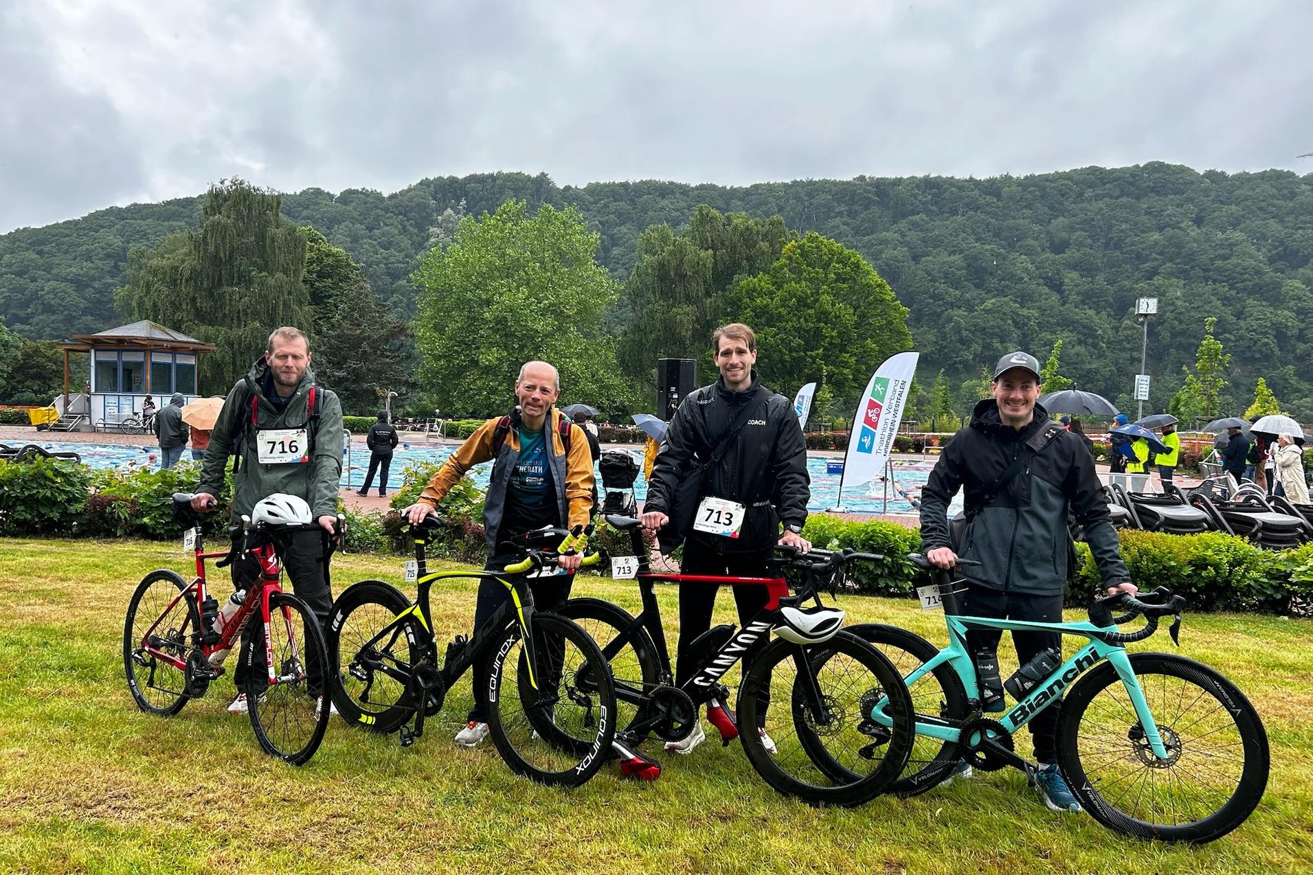 Vier Männer mit Fahrrädern stehen auf einer Wiese, im Hintergrund eine Schwimmhalle und grüne Hügel unter bewölktem Himmel.