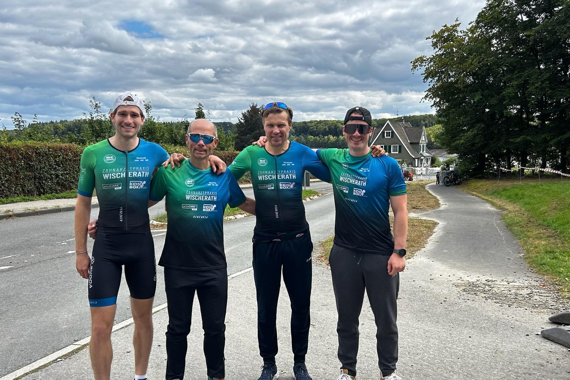 Vier Männer in Sportbekleidung stehen lächelnd an einer Straße, umgeben von Bäumen und Häusern.
