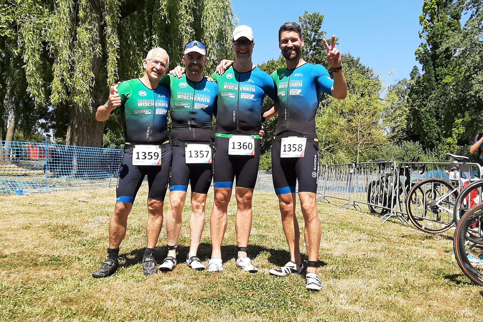 Vier Sportler in Wettkampfanzügen posieren lächelnd auf einer Wiese, umgeben von Fahrrädern und Absperrgittern.