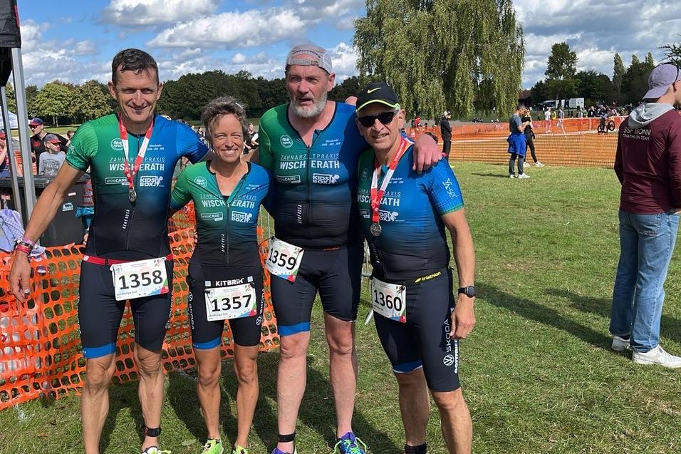 Vier Triathleten in Wettkampfanzügen steht in einem Gartenbereich nach dem Wettkampf, im Hintergrund Zuschauer und Zäune.