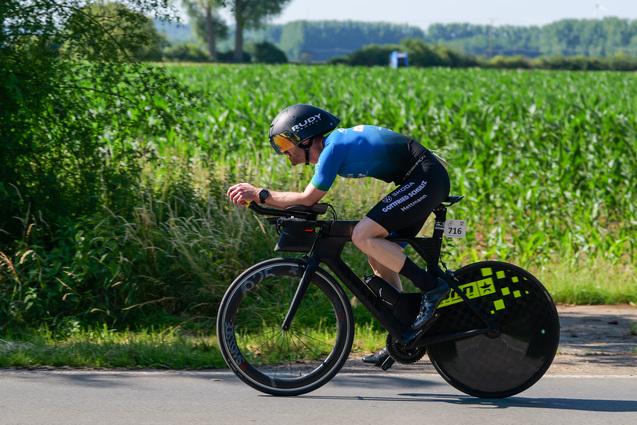 Radfahrer in aerodynamischer Position auf einem Zeitfahrrad, unterwegs auf einer Landstraße neben einem Getreidefeld.