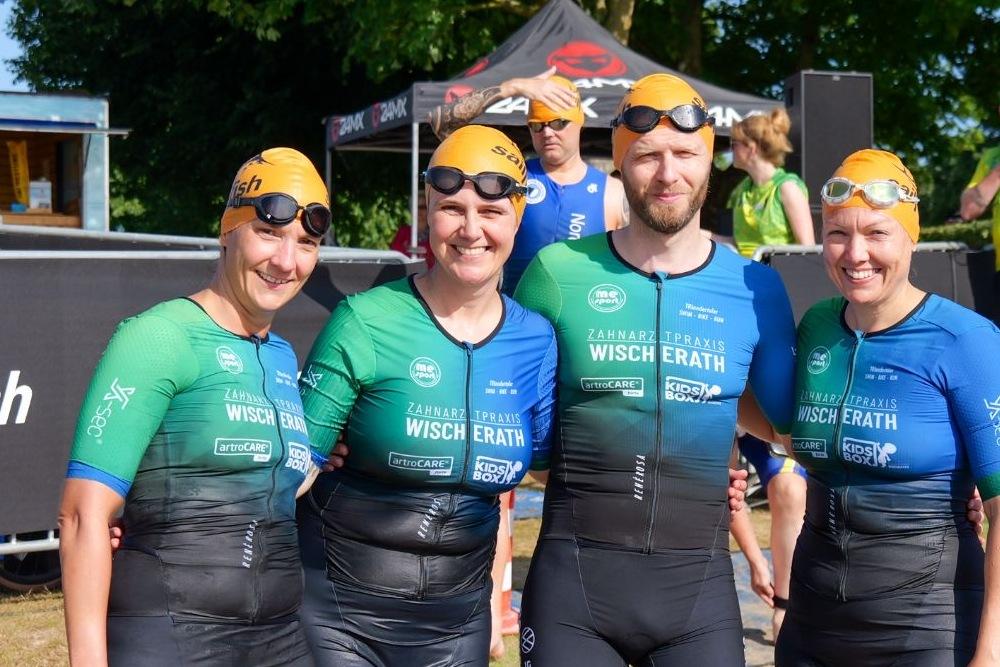 Vier Schwimmer in Neoprenanzügen und Badekappen stehen lächelnd am Ufer eines Wassersports-Events.