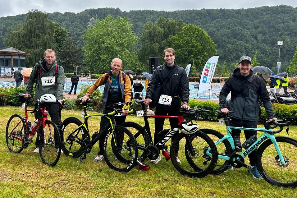Vier Männer mit Fahrrädern stehen auf einer Wiese, im Hintergrund eine Schwimmhalle und grüne Hügel unter bewölktem Himmel.