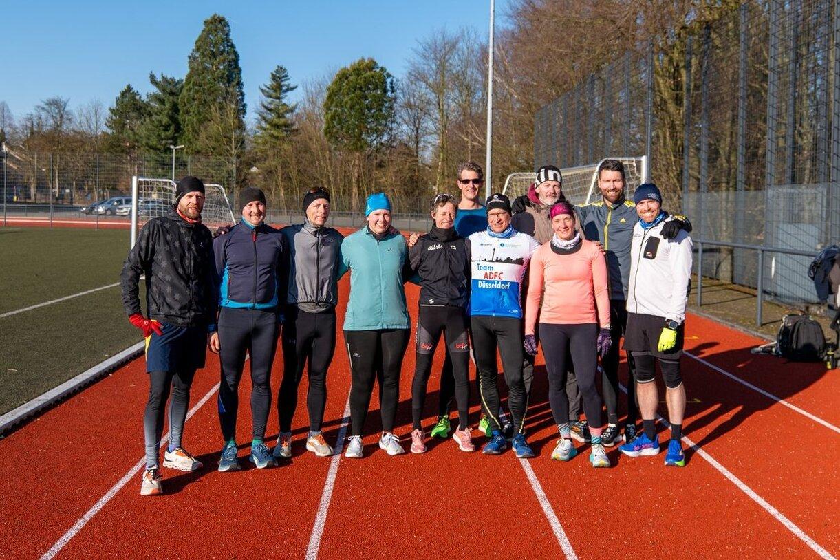 Gruppe von zehn L&auml;ufern auf einer roten Laufbahn, umgeben von B&auml;umen und einem Sportplatz im Hintergrund.