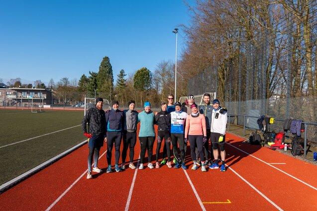 Gruppe von zehn Läufern auf einer roten Laufbahn, umgeben von Bäumen und einem Sportplatz im Hintergrund.