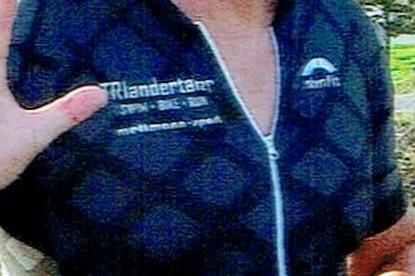 Lächelnder Teilnehmer eines Laufs in Sportkleidung mit Startnummer, joggt auf einem Weg mit Bäumen im Hintergrund.