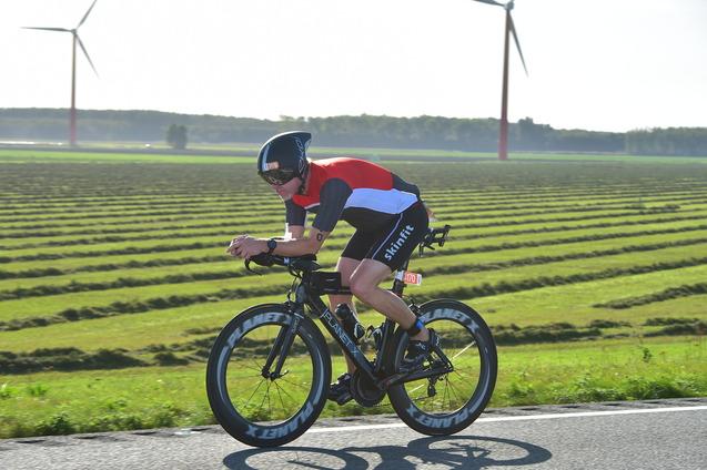 Radrennfahrer in sportlicher Kleidung fährt auf einer Landstraße, im Hintergrund Windkraftanlagen und gestreiftes Feld.