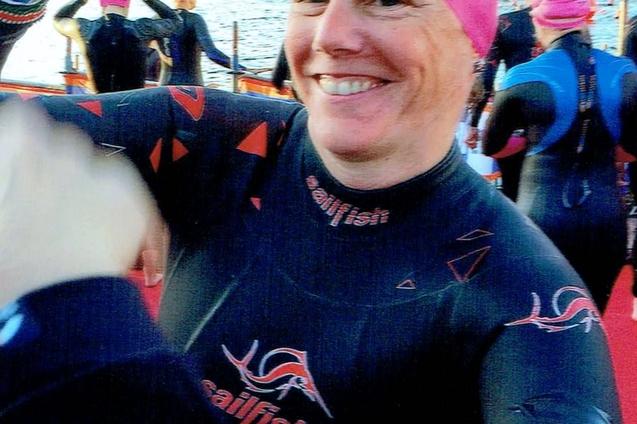 Lächelnde Schwimmerin in einem schwarzen Neoprenanzug und roter Badekappe, umgeben von weiteren Schwimmern am Wasser.