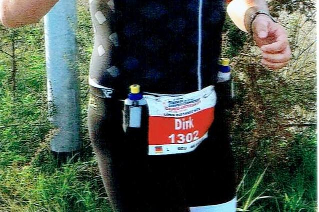 Lächelnder Teilnehmer eines Laufs in Sportkleidung mit Startnummer, joggt auf einem Weg mit Bäumen im Hintergrund.