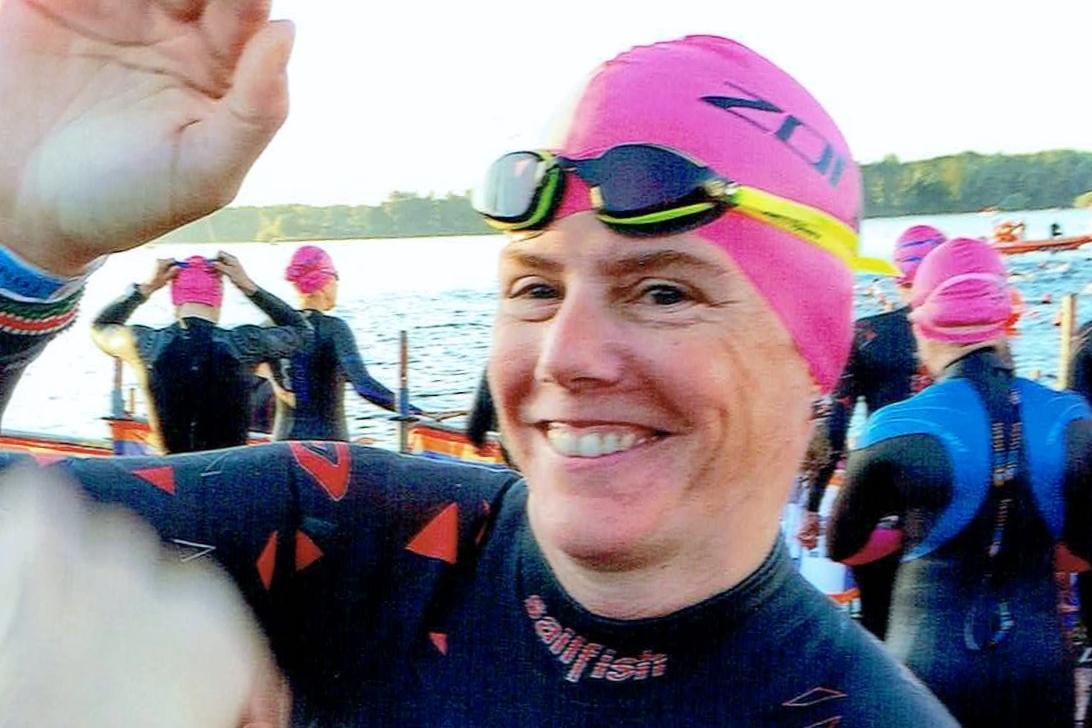 L&auml;chelnde Schwimmerin in einem schwarzen Neoprenanzug und roter Badekappe, umgeben von weiteren Schwimmern am Wasser.