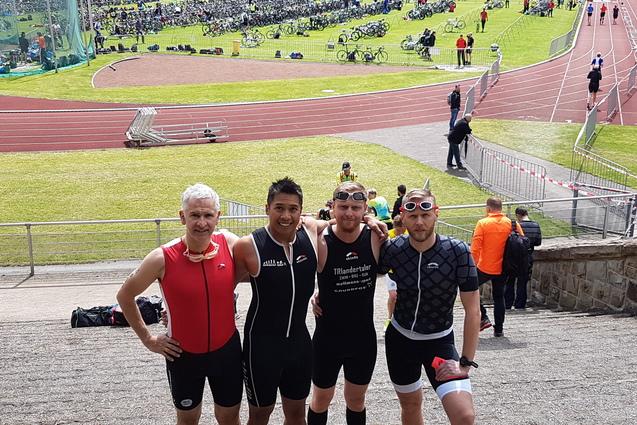Vier Triathleten in Sportkleidung posieren l&auml;chelnd vor einer Sportveranstaltung mit Radfahrern im Hintergrund.