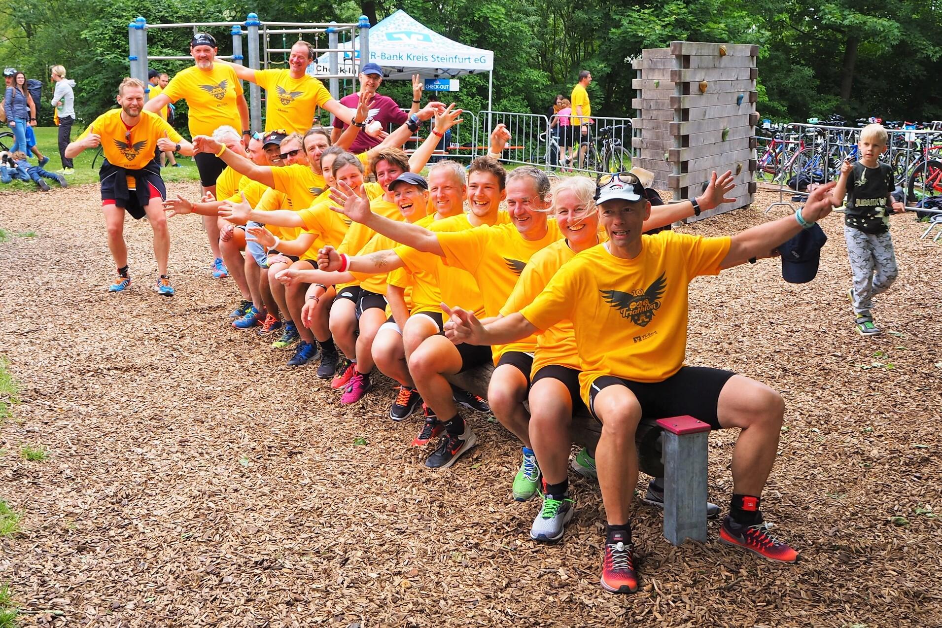 Gruppenselfie von Sportlern in gelben T-Shirts, die fröhlich auf einer Bank sitzen und die Arme heben.