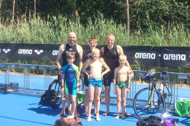 Gruppenfoto von drei Erwachsenen und vier Kindern in Sportkleidung neben Fahrrädern auf einer blauen Fläche.