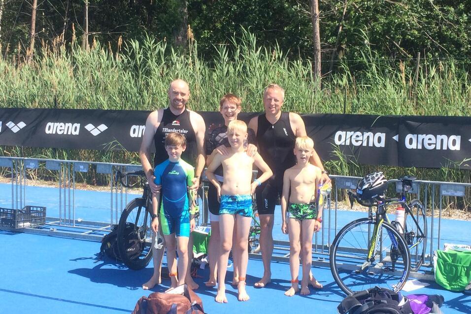 Gruppenfoto von drei Erwachsenen und vier Kindern in Sportkleidung neben Fahrrädern auf einer blauen Fläche.