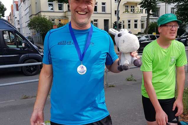 Lachender Mann in blauer Sportkleidung hält ein Stofftier und eine Tasche, im Hintergrund sind Bäume und Gebäude.